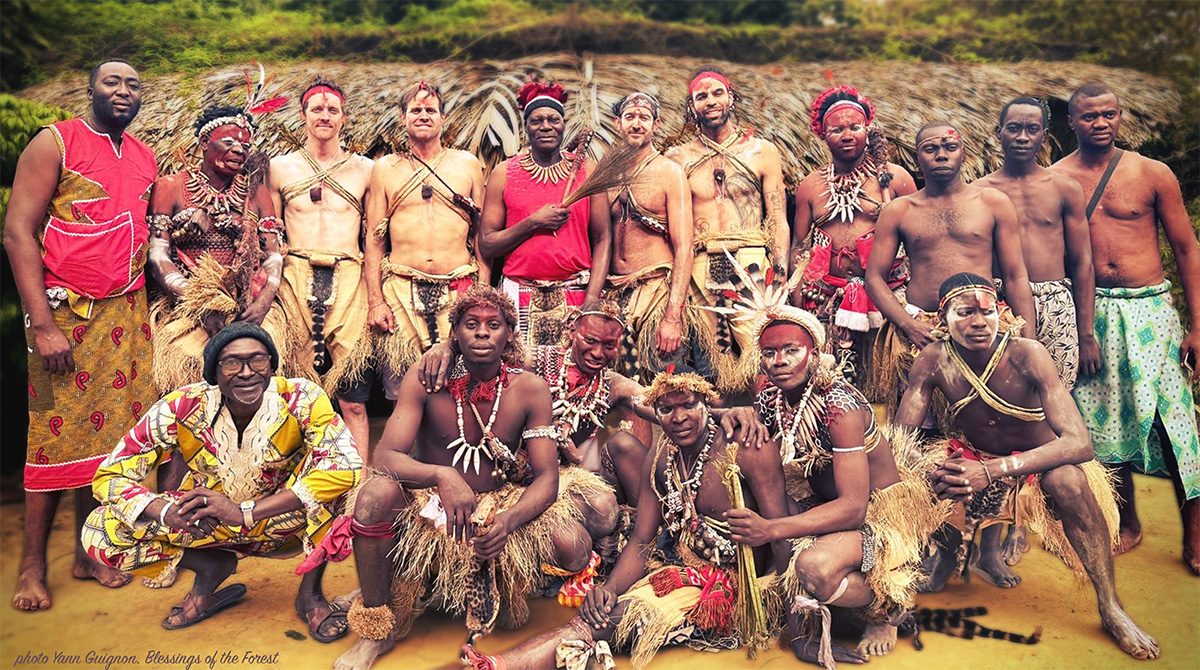 A large group of Bwiti initiates and visitors in ceremonial dress gathered outside a thatched temple in Gabon.