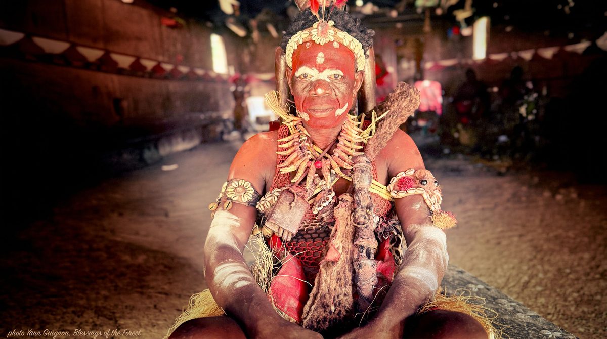 A Bwiti elder in full ceremonial dress and face paint, seated inside a temple in Gabon.