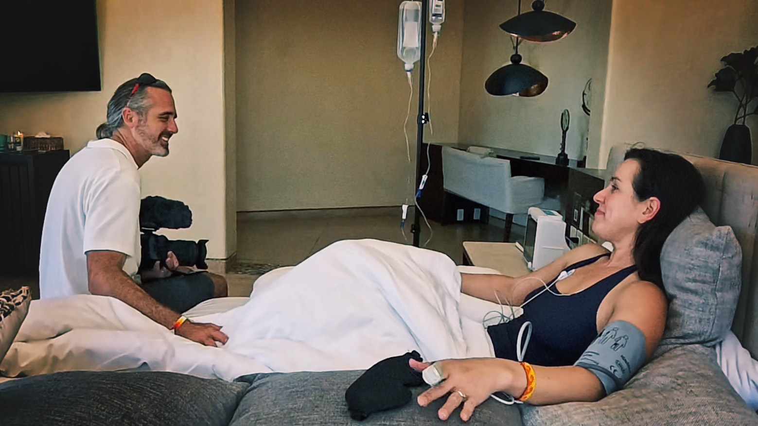 A patient resting on a monitored bed at an ibogaine clinic while a practitioner sits alongside.